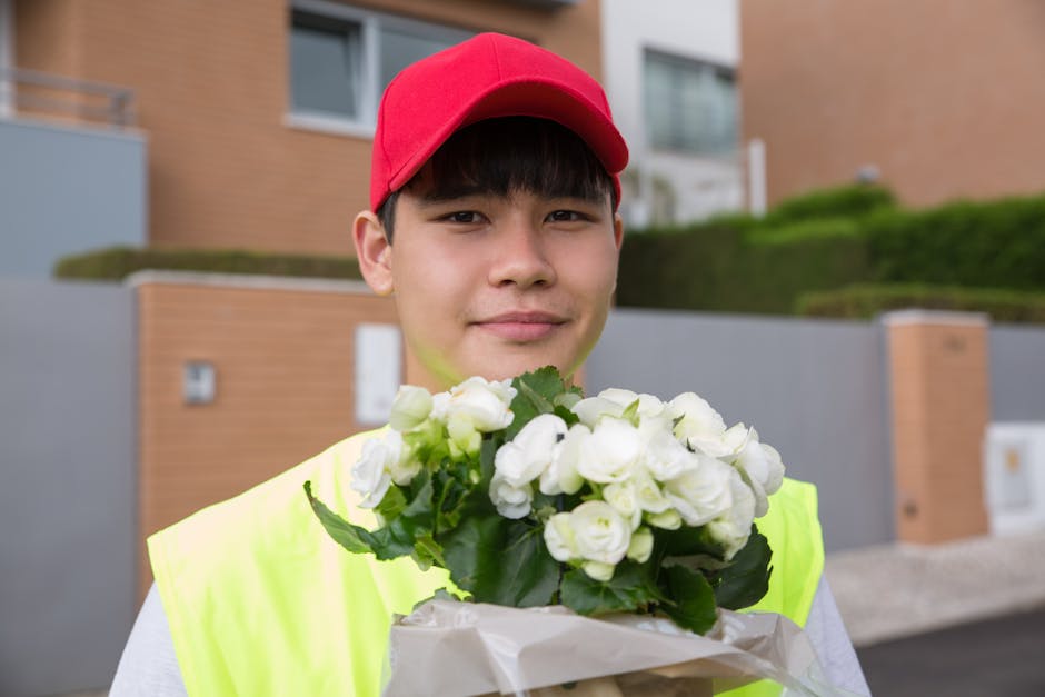 A young male delivery person with fair skin, wearing a red cap and a high-visibility yellow vest, is standing outdoors in a residential area with modern brick buildings and trimmed hedges in the backg