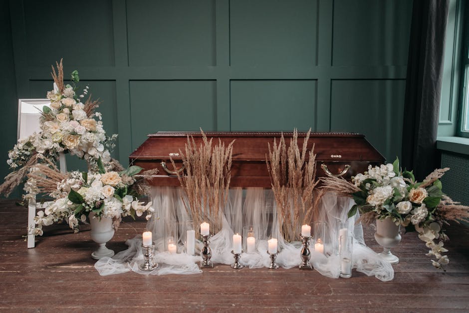 A somber floral display arranged on a wooden surface in front of a green paneled wall, featuring two large white vases filled with soft cream and white roses, hydrangeas, and greenery, complemented by