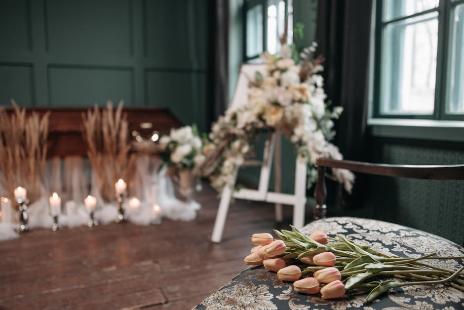 A floral arrangement setup with a bouquet of pale pink tulips with green stems and leaves resting on a decorative upholstered chair in the foreground. In the background, a large, elegant floral tribut