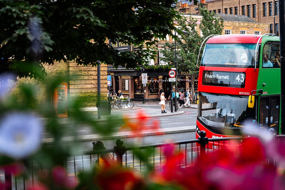 A busy street scene in Friern Barnet with a red and green double-decker bus displaying the route number 43 and the destination 'Friern Barnet.' In the foreground, out-of-focus vibrant pink, red, and w