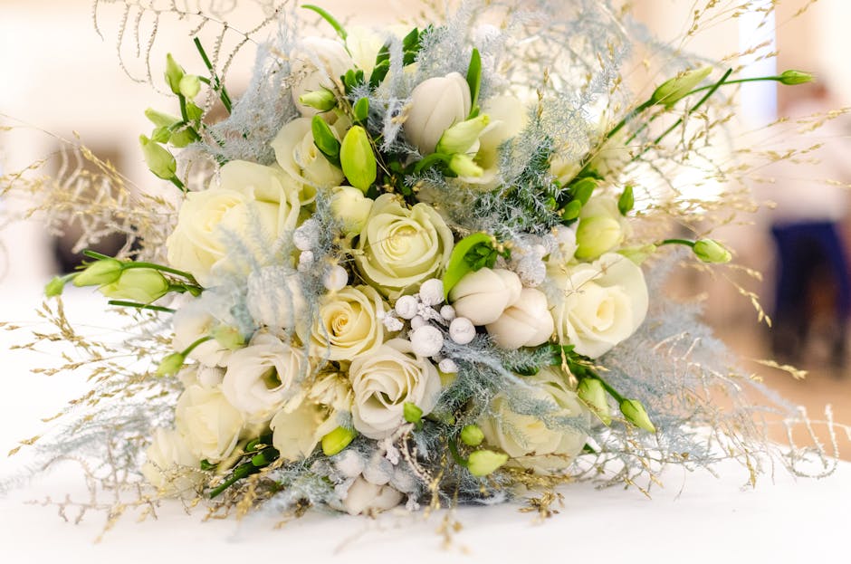 A floral arrangement featuring a bouquet of white roses, tulips, and lisianthus with delicate green buds, accented by silvery foliage and frosted decorative elements. The bouquet is placed on a white