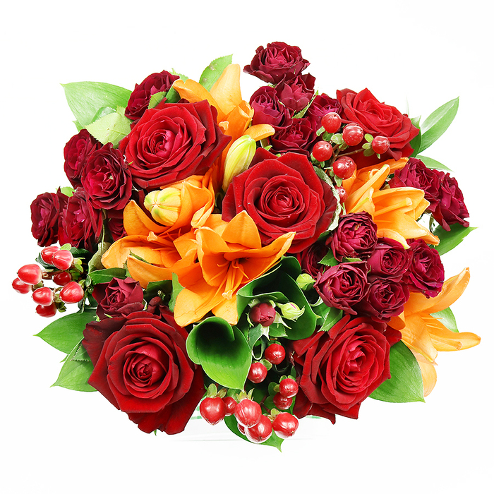 Captured overhead on a neutral white ground, this lavish bouquet reads like a small jewel - intentionally dense, circular and composed to show every texture and tone. Several generously sized, fully opened deep red roses anchor the arrangement with layered, velveteen petals that fold inward toward intricate centres; their classic fragrance suggests long-stemmed romance. Bright, fiery orange Asiatic lilies slice through the red with trumpet-shaped blooms and prominent stamens, some blossoms fully expanded and freckled while others hold as elegant buds, creating a feeling of motion and freshness. Interwoven are clusters of dark burgundy spray roses-tight, luxurious miniatures that introduce shadowed richness and compact volume. Scattered among the blooms, glossy hypericum berries in a true ruby red add contrast both visually and texturally, their spherical shine picking up the soft studio light. Lush green foliage-broad, slightly glossy leaves reminiscent of ruscus or salal-fans out and between the flowers to give breathing space and botanical structure. The lighting is soft and even, allowing the eye to trace velvety petal edges, the stamens' fine filaments, and the subtle sheen on berries without distraction. This composition evokes warmth, celebration and intimate joy, perfect as a gift for anniversaries or a striking centrepiece in a Friern Barnet home near Colney Hatch Lane, Friary Park or the shops along Friern Barnet Road. As a florist's piece, it balances opulence with approachability-a bouquet designed to be both seen and savoured.