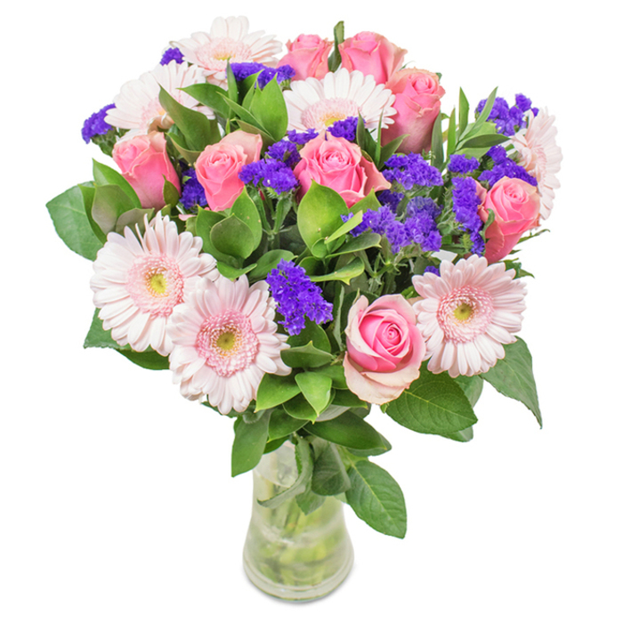 An exuberant floral bouquet, carefully curated and presented in a clear glass vase, sits against a pure white backdrop-a modern classic crafted for Friern Barnet's vibrant community. At the heart of the arrangement are around eight pink roses, each at varying stages of unfurling, their spiral petals exuding both elegance and a sense of cherished tradition. Surrounding these, several soft pink gerbera daisies-distinguished by their generous, round blossoms and lighter, almost snowy centres-bring a gentle innocence to the composition. Interspersed are rich purple statice clusters, providing vivid colour contrast and tactile visual interest, recalling the diverse flora of Princess Park. Lush, deep green foliage wraps the ensemble in a mantle of freshness, evoking the leafy thoroughfares and private gardens found throughout Friern Barnet. The transparent glass vase, partially filled with water, allows glimpses of intersecting green stems, reinforcing the bouquet's freshly gathered, natural feel. This inviting arrangement, perfect for birthdays, anniversaries, or as a daily token of affection, delivers a message of celebration and heartfelt connection, grounded in the local colour and warmth of Friern Barnet's neighbourhood spirit.