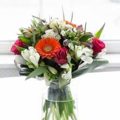 This exuberant bouquet, arranged with the artistry of Friern Barnet's finest florists, captures a tapestry of colours and textures in a single, flourishing composition. Springing from a graceful clear glass vase, the display features prominent gerbera daisies-two ablaze in warm, spirited orange and two in the gentlest of peach, their soft petals surrounding velvety brown centres reminiscent of late afternoon sunlight flickering through the trees at Friary Park. Nestled among them are opulent, tightly furled fuchsia roses, adding a lavish touch of romance and depth that calls to mind the neighbourhood's centuries-old charm. Clusters of pure white alstroemeria and dainty white spray roses glide throughout the arrangement, infusing it with brightness, airiness, and a senses-awakening freshness. Lighter pink blossoms whisper of new beginnings, while abundant, glossy green leaves-some artistically folded into broad curves, others pointed and sprightly-wrap protectively around the stems, echoing the leafy calm of local avenues. Wispy sprigs weave between the petals, building a bouquet that feels at once structured and abundantly free. The soft light pouring across the scene enhances the vibrancy of every element, spotlighting the expertise and attention to detail that defines Friern Barnet floristry. Ideal for thoughtful celebrations, housewarmings, or simply sharing joy with someone special, this bouquet is a glowing tribute to local beauty and heartfelt connection.
